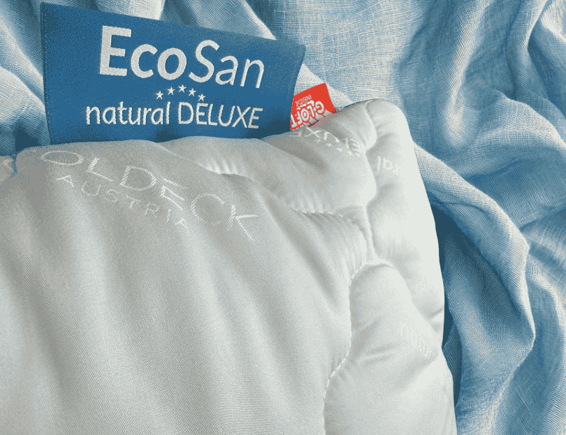 Nahaufnahme eines gesteppten Kissens mit eingesticktem „Goldeck Austria“-Schriftzug und sichtbaren Labels „EcoSan natural DELUXE“ sowie „G-LOFT INSIDE“, drapiert auf weichem Stoff.